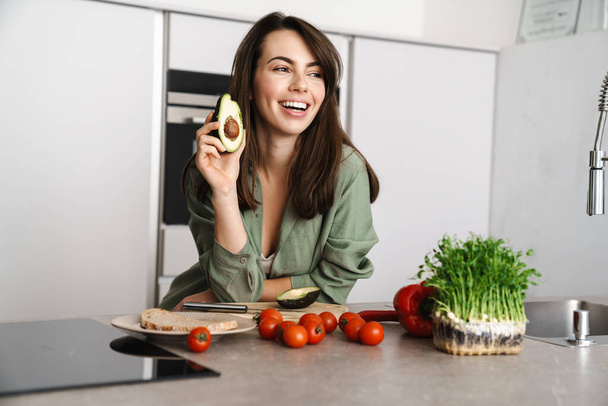 Cheerful beautiful woman showing avocado while making salad at home kitchen - Foto, afbeelding