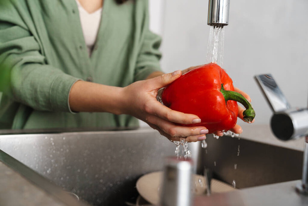 Caucasian young woman washing bell pepper at home kitchen - 写真・画像