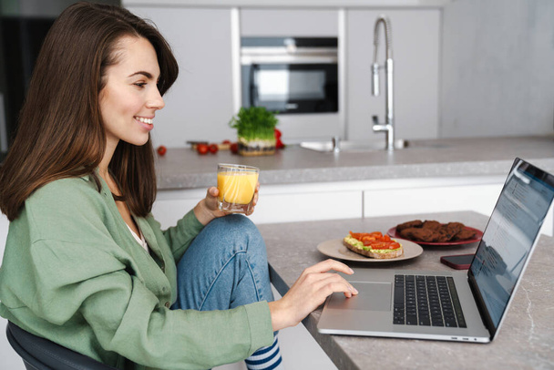Attractive young woman working on laptop while sitting in the kitchen at home - Zdjęcie, obraz