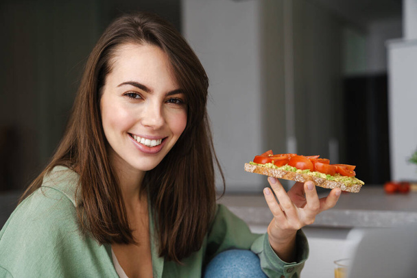 Young happy woman smiling while eating avocado toast at home kitchen - Fotoğraf, Görsel