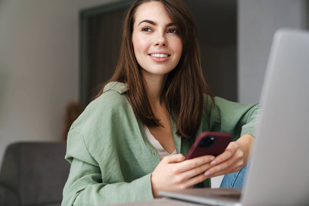 Cheerful beautiful woman using mobile phone while working with laptop at home - Photo, Image