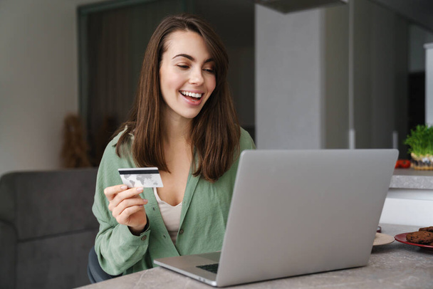 Cheerful nice woman using credit card while working with laptop at home - Φωτογραφία, εικόνα