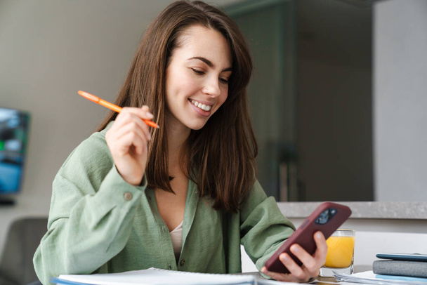 Young smiling woman using mobile phone while working with paper at home kitchen - Photo, image