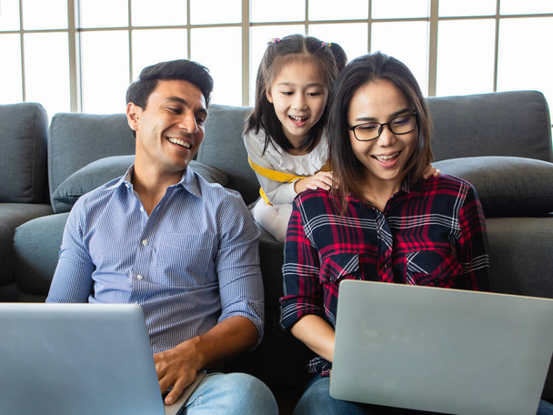 Három különböző családtag, kaukázusi apa, ázsiai anya és kislánya együtt ülnek a nappaliban, és 3 laptopos notebook számítógépet használnak. Ötletek az otthoni munkavégzéshez. - Fotó, kép