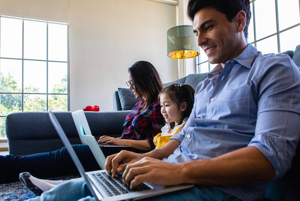 Drei Familienmitglieder, ein kaukasischer Vater, eine asiatische Mutter und eine kleine Halbtochter sitzen zusammen im Wohnzimmer des Hauses und bedienen drei Laptops. Idee für die Arbeit zu Hause. - Foto, Bild