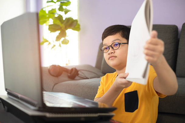 若くかわいい10代のアジア系の少年は、自宅のリビングルームでノートパソコンを使用してオンライン学習中に教師のための宿題を示す眼鏡をかけていました。距離教育思想. - 写真・画像