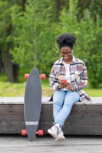 Happy african skater girl texting online in smartphone app sit with longboard on bench in skate park - Фото, изображение