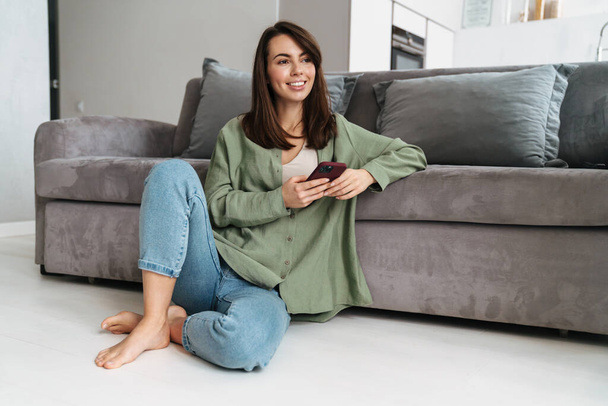 Smiling attractive woman using mobile phone while sitting on a floor at home - Photo, Image