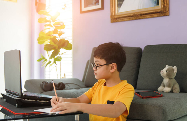 Jong jongetje met een bril in de buurt van een bank in de woonkamer thuis en huiswerk maken tijdens de les leren via laptop notebook computer. - Foto, afbeelding