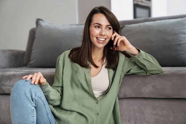 Cheerful beautiful woman talking on mobile phone while sitting on floor at home - Photo, image