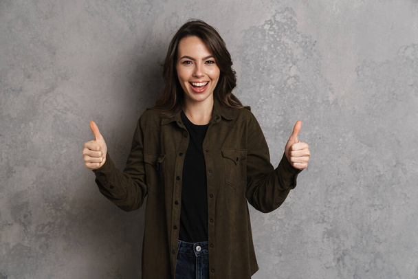 Портрет счастливой улыбающейся женщины в повседневной одежде, стоящей изолированно на сером фоне, показывая большие пальцы вверх - Фото, изображение
