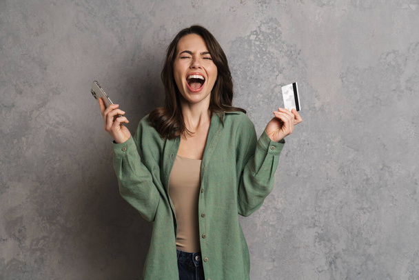 Excited girl screaming while posing with cellphone and credit card isolated over grey wall - Fotó, kép