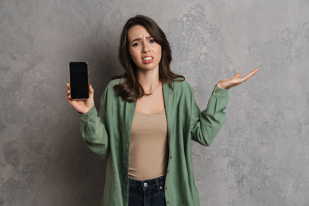 Összezavarodott lány kezében copyspace miközben mutatja mobiltelefon elszigetelt szürke fal - Fotó, kép