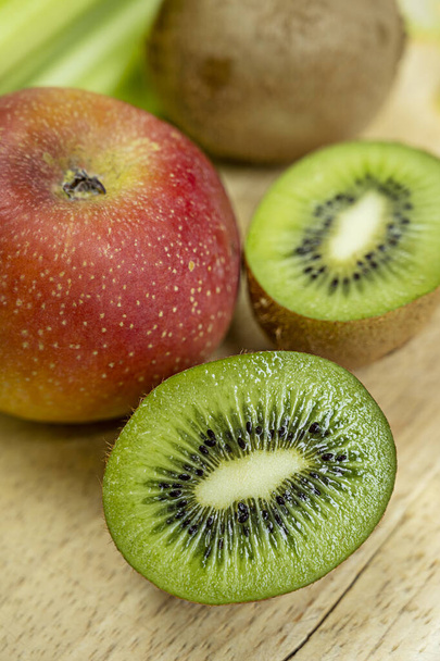 Close-up of kiwi cut in half and more kiwis in the background with red apple on light wooden background - Foto, Imagem