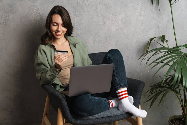 Smiling nice girl using laptop and credit card while sitting in chair indoors - Photo, image