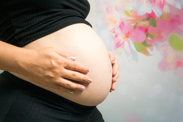 Pancia di una donna bianca incinta con entrambe le mani posizionate intorno al bambino indossando un vestito nero su uno sfondo colorato e fiorito. Menzionare le nuove madri e le sfide della gravidanza. - Foto, immagini