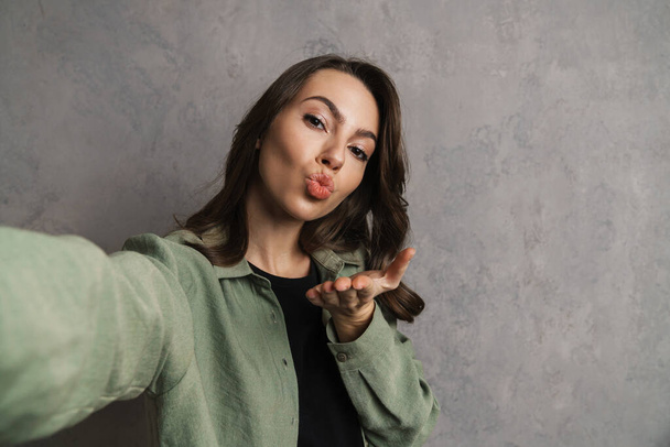 Happy beautiful girl blowing air kiss while taking selfie photo isolated over grey wall - Foto, imagen