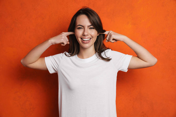 Happy beautiful brunette girl smiling while plugging her ears isolated over orange wall - Zdjęcie, obraz