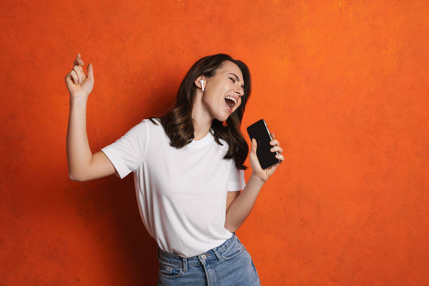 Happy girl in earphones singing and dancing while using cellphone isolated over orange wall - Fotoğraf, Görsel