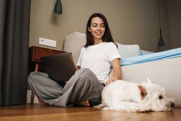 Happy white woman using laptop and stroking her while sitting on floor in home bedroom - Photo, Image