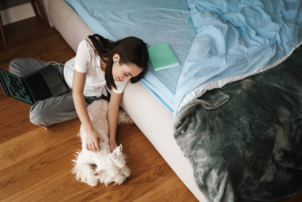 Happy young brunette woman stydying in bedroom, sitting on the floor with her dog and laptop computer - 写真・画像
