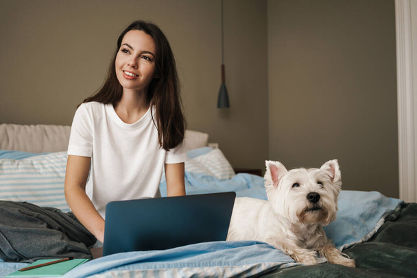 Happy young brunette woman stydying in bedroom, sitting on bed with her dog and laptop computer - Foto, imagen