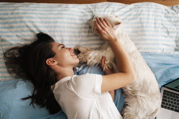 Happy white woman stroking her dog while lying on bed at home - Фото, изображение