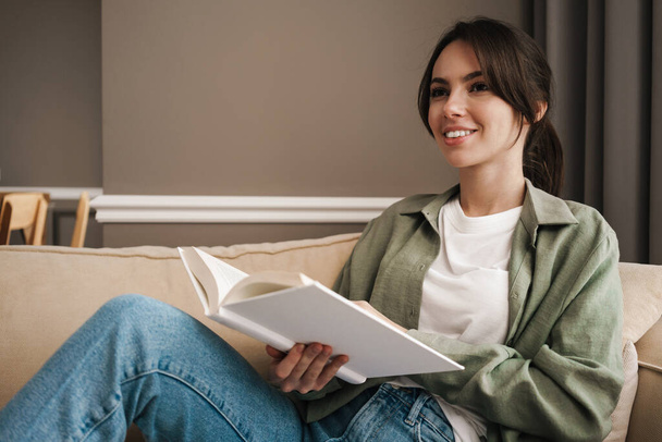 Retrato de una joven feliz sentada en un sofá con libro - Foto, Imagen