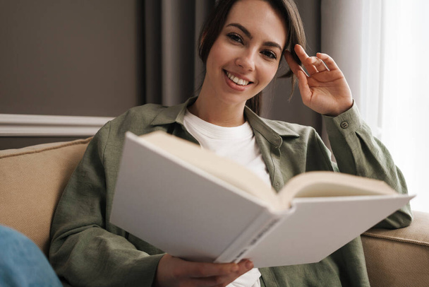 Portrait of a happy young woman sitting on couch with book - Foto, imagen