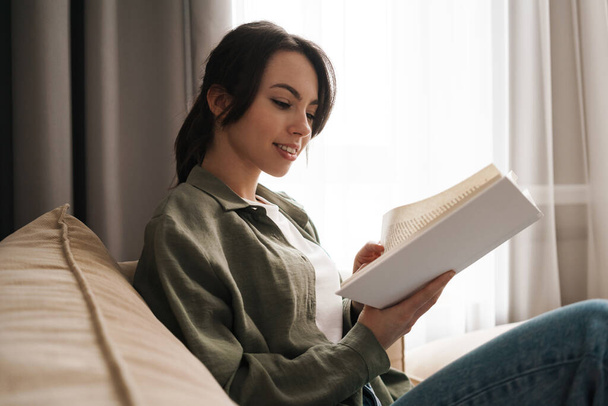 Portrait of a happy young woman sitting on couch with book - Photo, Image