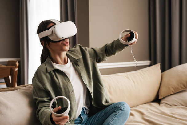 Excite white woman playing online game with vr glasses and controllers on sofa at home - Zdjęcie, obraz