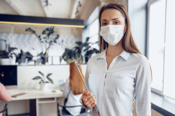 Young businesswoman in face mask standing in office and holding clipboard with documents - Фото, изображение
