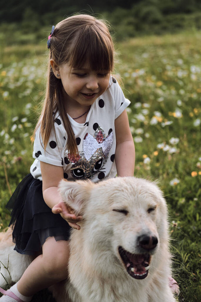 Słodka biała dziewczynka siedzi na dużym białym psie i uśmiecha się. Najlepszymi przyjaciółmi są dzieci i psy. Połowa rasy biały owczarek szwajcarski w rumianku pole leży na uśmiechy. - Zdjęcie, obraz