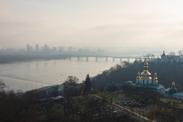 Una fitta nebbia sul fiume con un ponte e una città sull'altra sponda. Chiesa ortodossa e cortile del monastero - Foto, immagini