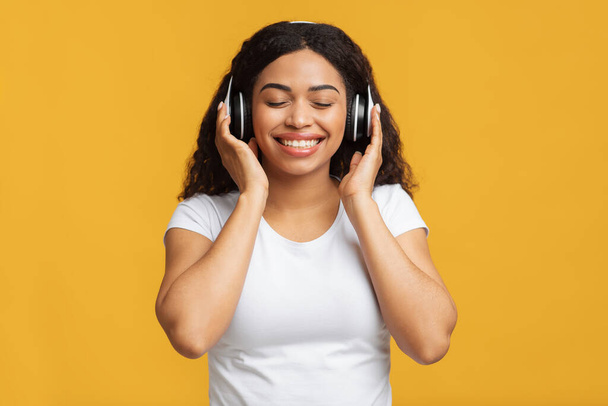 Дама с закрытыми глазами слушает музыку на желтом фоне, используя беспроводные наушники - Фото, изображение