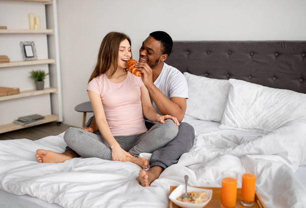 Longitud completa de tipo negro alimentando croissant fresco sabroso a su novia, sentado en la cama en casa, espacio de copia - Foto, Imagen