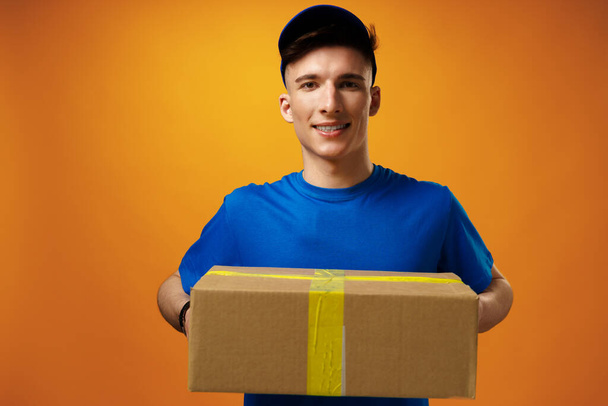 Young handsome delivery man holding parcel cardboard box against yellow background - Foto, Bild
