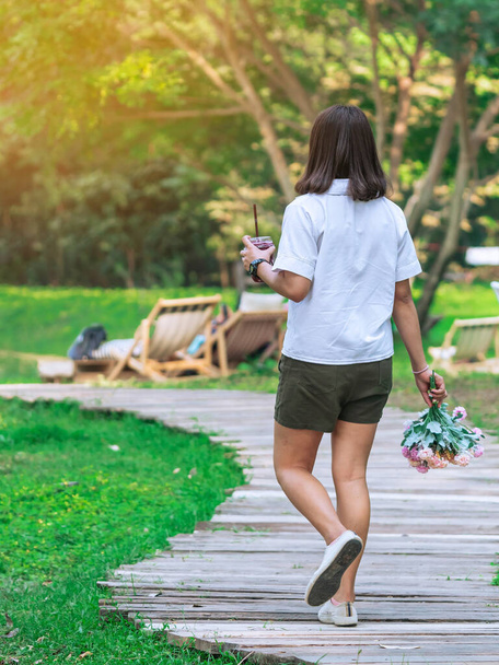 Вид на азиатскую женщину с напитками и букет ходить в одиночку по дорожке через зеленый сад. Женщина отдыхает одна в парке. Люди проводят время на улице в зеленой природе. Наслаждение природой на открытом воздухе. - Фото, изображение