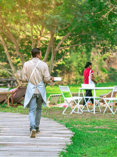 Back view of Asian male waiter carry food and beverages along garden paths to serve customers while relaxing in open-air restaurant. People spending time outside in green nature. Enjoying outdoors. - Photo, Image