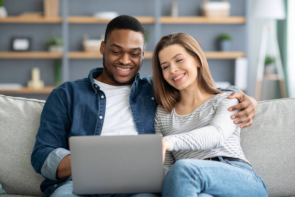 Ευτυχισμένο νεαρό διαφυλετικό ζευγάρι με φορητό υπολογιστή στο σπίτι ψώνια σε απευθείας σύνδεση - Φωτογραφία, εικόνα