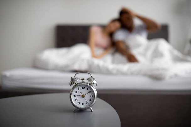 Verschwommenes multiethnisches Paar, das morgens verschlafen wird, zu Hause auf dem Bett sitzt, spät zur Arbeit geht, sich auf den Wecker konzentriert - Foto, Bild