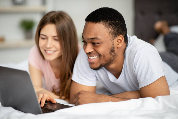 多人種のカップルを愛情を込めて自宅で一緒に時間を過ごします,ベッドの上でラップトップを使用して - 写真・画像