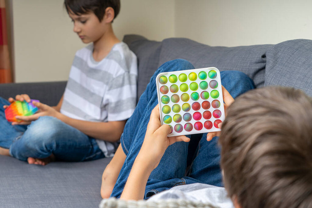 Nastoletni chłopcy bawią się tęczowymi zabawkami w domu. Push pop-it fidgeting gra pomaga złagodzić stres, lęk, autyzm, zapewnić wrażenia sensoryczne i dotykowe dla dzieci. - Zdjęcie, obraz