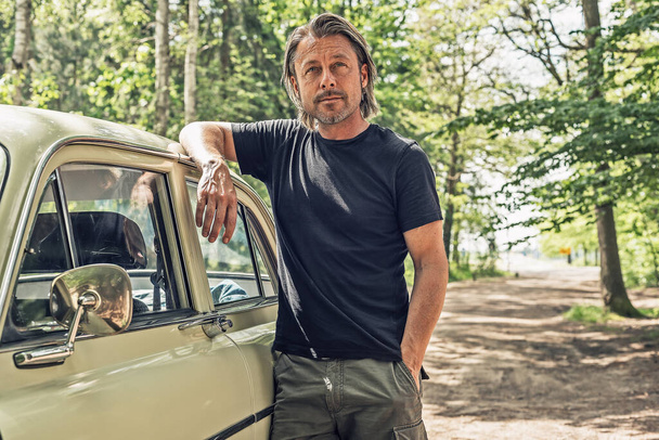 Man in a black t-shirt standing by a classic car on a forest dirt road. - Fotografie, Obrázek