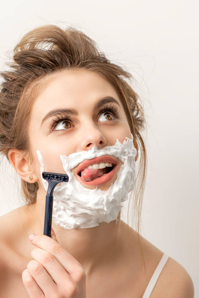 Красива молода біла усміхнена жінка голиться обличчям з бритвою, що стирчить язиком, дивлячись на білий фон
 - Фото, зображення