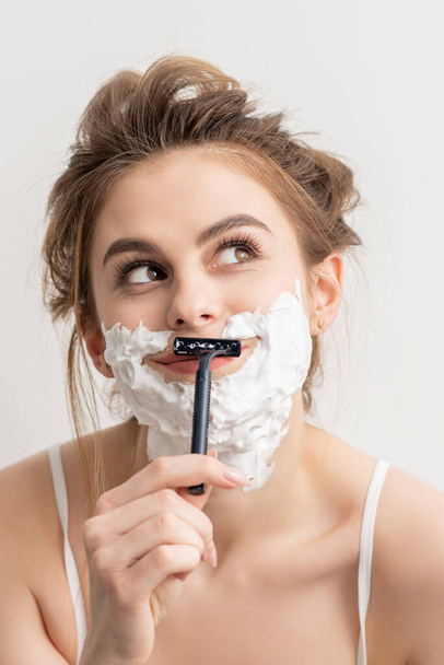 Beautiful young caucasian smiling woman shaving her face with razor looking up on white background - Photo, Image
