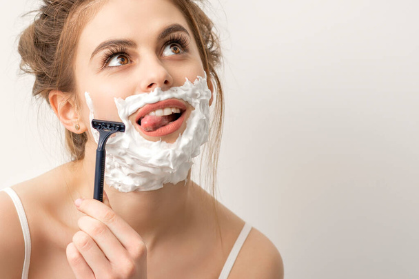 Красива молода біла усміхнена жінка голиться обличчям з бритвою, що стирчить язиком, дивлячись на білий фон
 - Фото, зображення