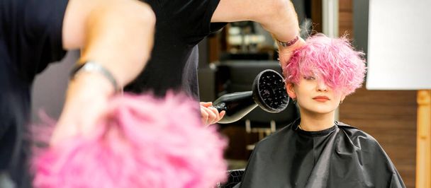 Un profesional de peluquería masculina secado elegante pelo rosa de la cliente femenina con un secador de pelo en un salón de belleza - Foto, Imagen