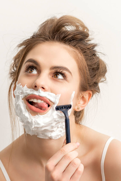 Beautiful young caucasian smiling woman shaving her face with razor sticking out tongue looking up on white background - Photo, Image