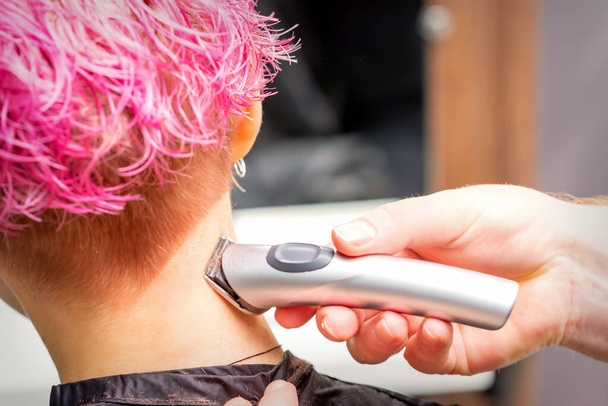 Вид ззаду рука перукаря голиться потилиця і шия з електричним тримером молодої кавказької жінки з коротким рожевим волоссям в салоні краси
 - Фото, зображення